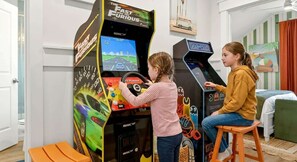 Zwei Spielautomaten für Rennspiele in einem Raum mit einem Bett und einer Wand mit Streifenmuster.