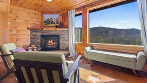 L’intérieur chaleureux d’un chalet avec une cheminée en pierre, des murs en bois, une grande fenêtre donnant sur les montagnes et des sièges confortables.