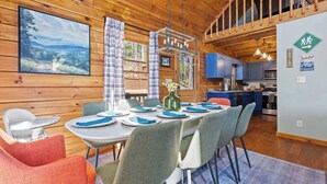 L’intérieur d’un chalet en bois comprend une salle à manger avec une table dressée pour six personnes, une cuisine aux armoires bleues et un grand tableau accroché au mur.