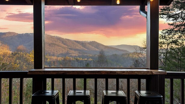 Une terrasse en bois avec trois tabourets noirs et une vue sur une chaîne de montagnes boisées au coucher du soleil.