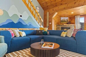 Ein gemütliches Wohnzimmer mit einem blauen Sofa, einem runden Holztisch, einem Kamin und einer Wandmalerei von Bergen.