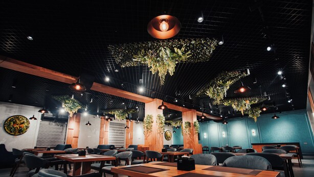 A modern restaurant interior with wooden tables, black chairs, hanging plants, and pendant lighting. - Bagishamol Hotel (Urgut)