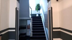 Un pasillo con piso a rayas blanco y negro, una puerta blanca al final y una escalera que sube a un piso superior.