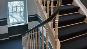 Una escalera con barandillas blancas y peldaños oscuros, que conduce a un rellano con un ventanal.