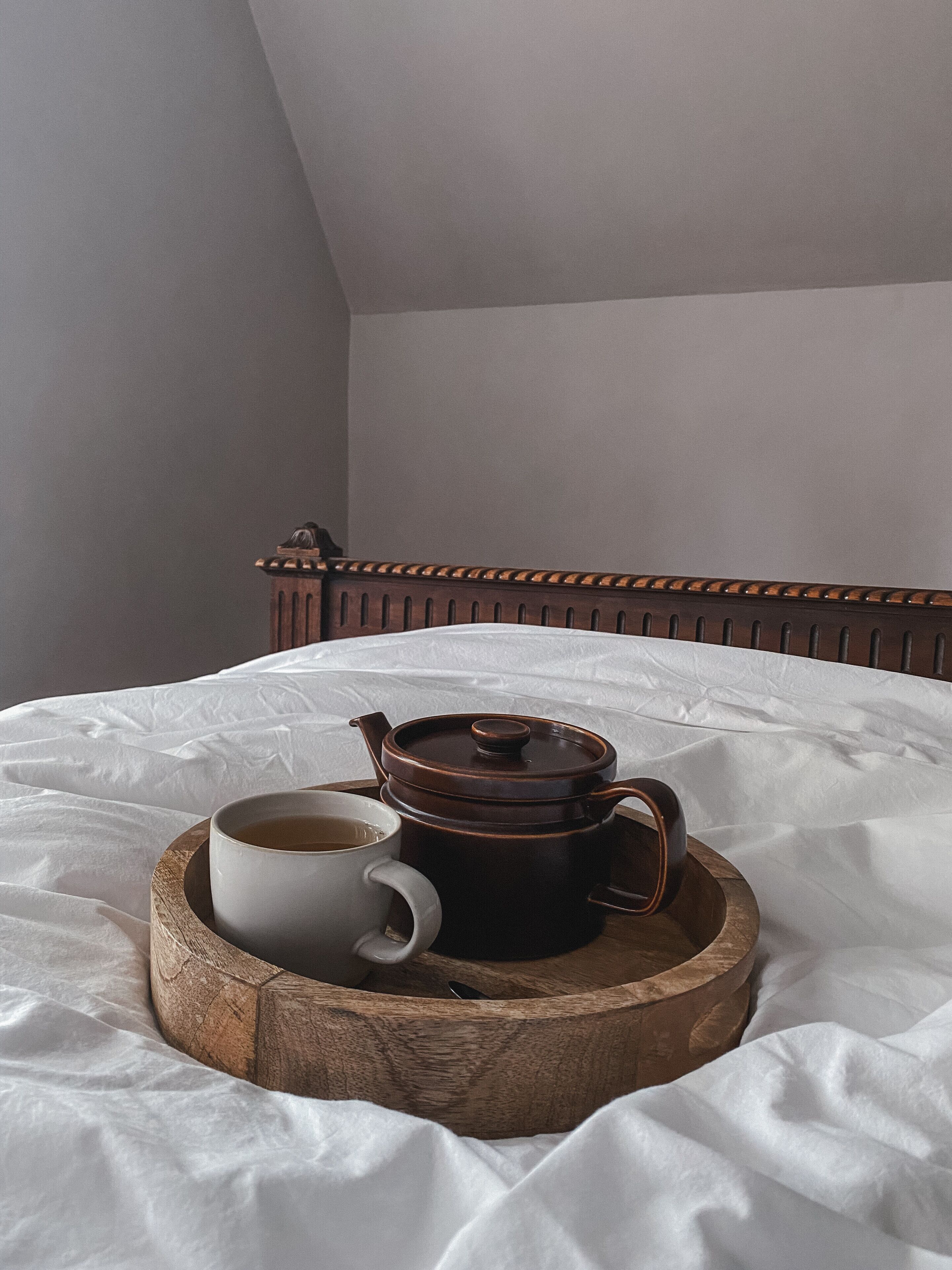 Un lit sur lequel repose un plateau en bois, portant une tasse blanche et un pot de couleur marron foncé. L’ensemble est placé devant un mur uni et sous un plafond en pente.