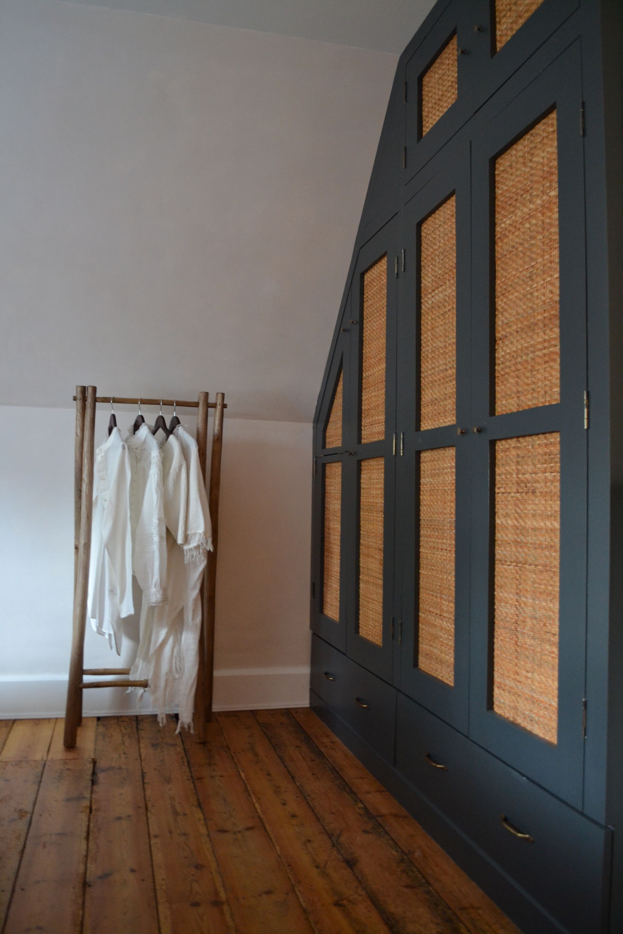 A room with a wooden floor, a clothing rack with white garments, and a dark blue wardrobe with woven panels.