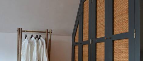 A room with a wooden floor, a clothing rack with white garments, and a dark blue wardrobe with woven panels.