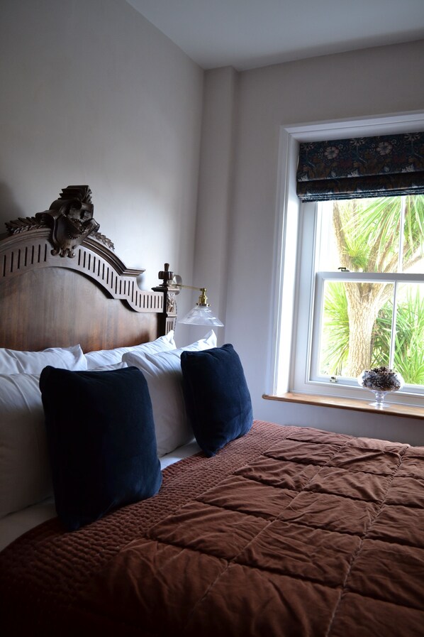 A bedroom with a wooden headboard, a brown quilted bedspread, two dark pillows, a bedside table with a lamp, and a window with a view of greenery. - Relaxing retreat in an 1880's Chapel development, coastlines & national parks. (Taunton)