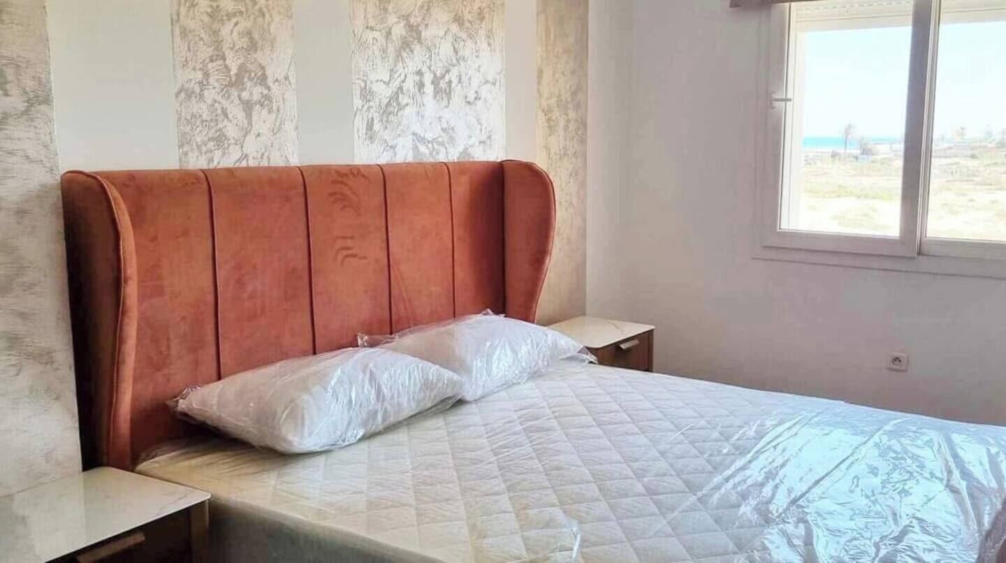Ein Schlafzimmer mit einem Bett, einem Holz-Kopfschild, zwei Nachttischen und einem Fenster mit Blick auf den Strand.