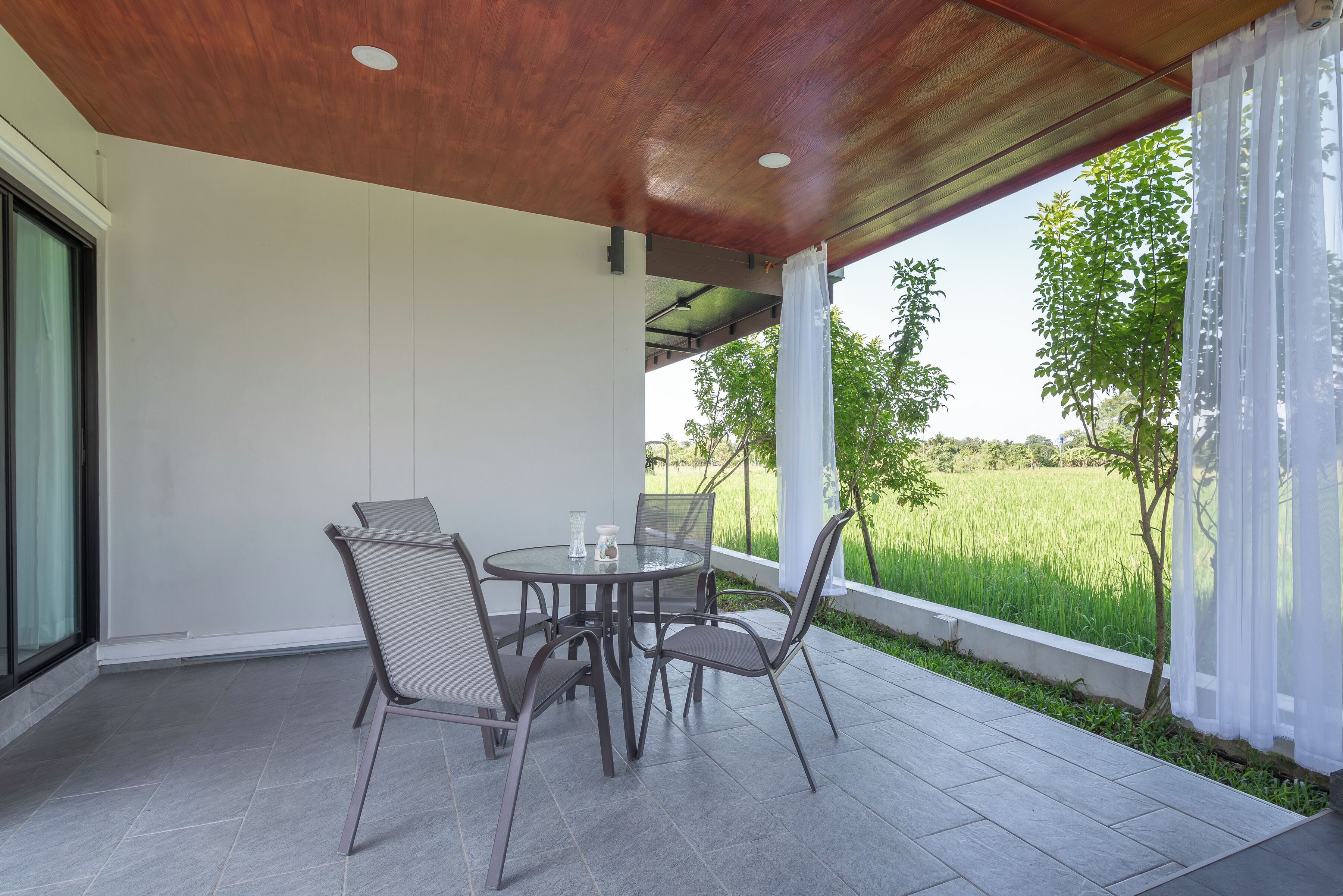 A covered patio with a table and chairs, a view of a green field, and a white wall.