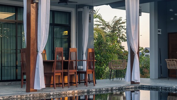 Un espace extérieur avec une piscine, une table et des chaises en bois, un coin salon abrité et une vue dégagée sur la végétation environnante.