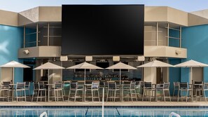 Zona de bar junto a la piscina con sillones de descanso, sombrillas, una pantalla grande y una barra llena de botellas.