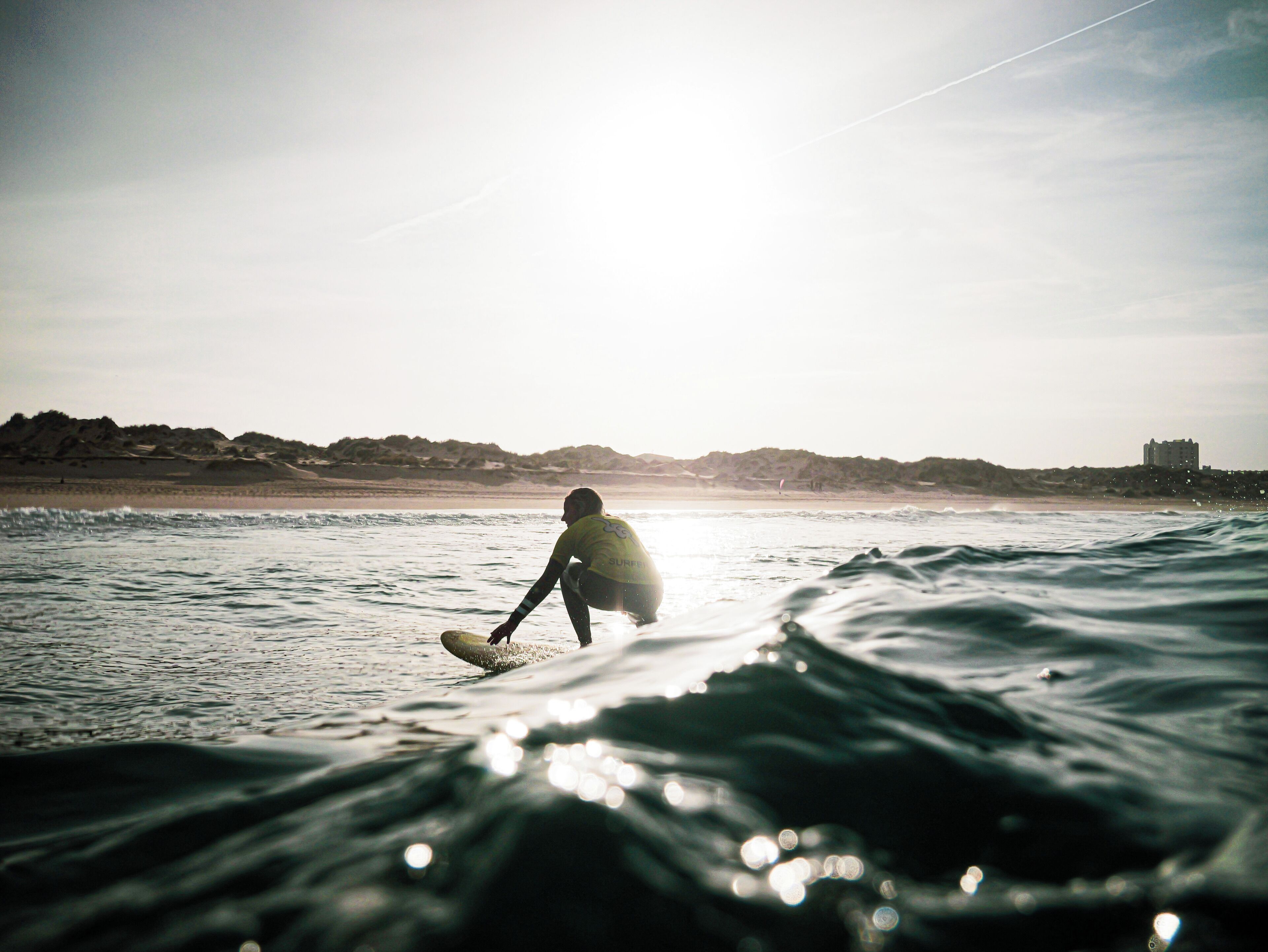 Ein Surfer auf einer Welle, im Hintergrund Küste und Gebäude.