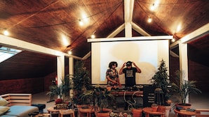 Een gezellige kamer met een houten plafond, een projectiescherm en een dj-installatie. Er staan verschillende plantenbakken met planten en een bank.
