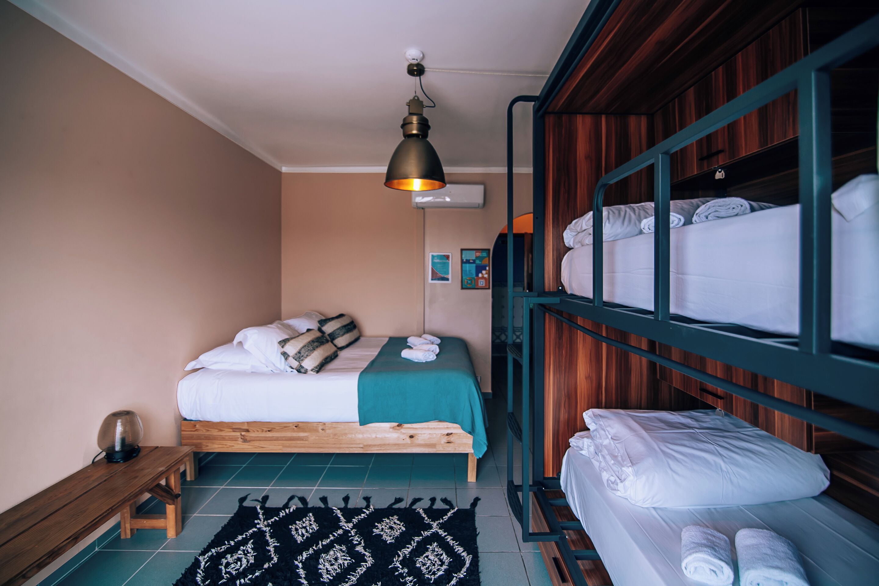 A bedroom with a single bed, a bench, a hanging light fixture, and a bunk bed with towels.