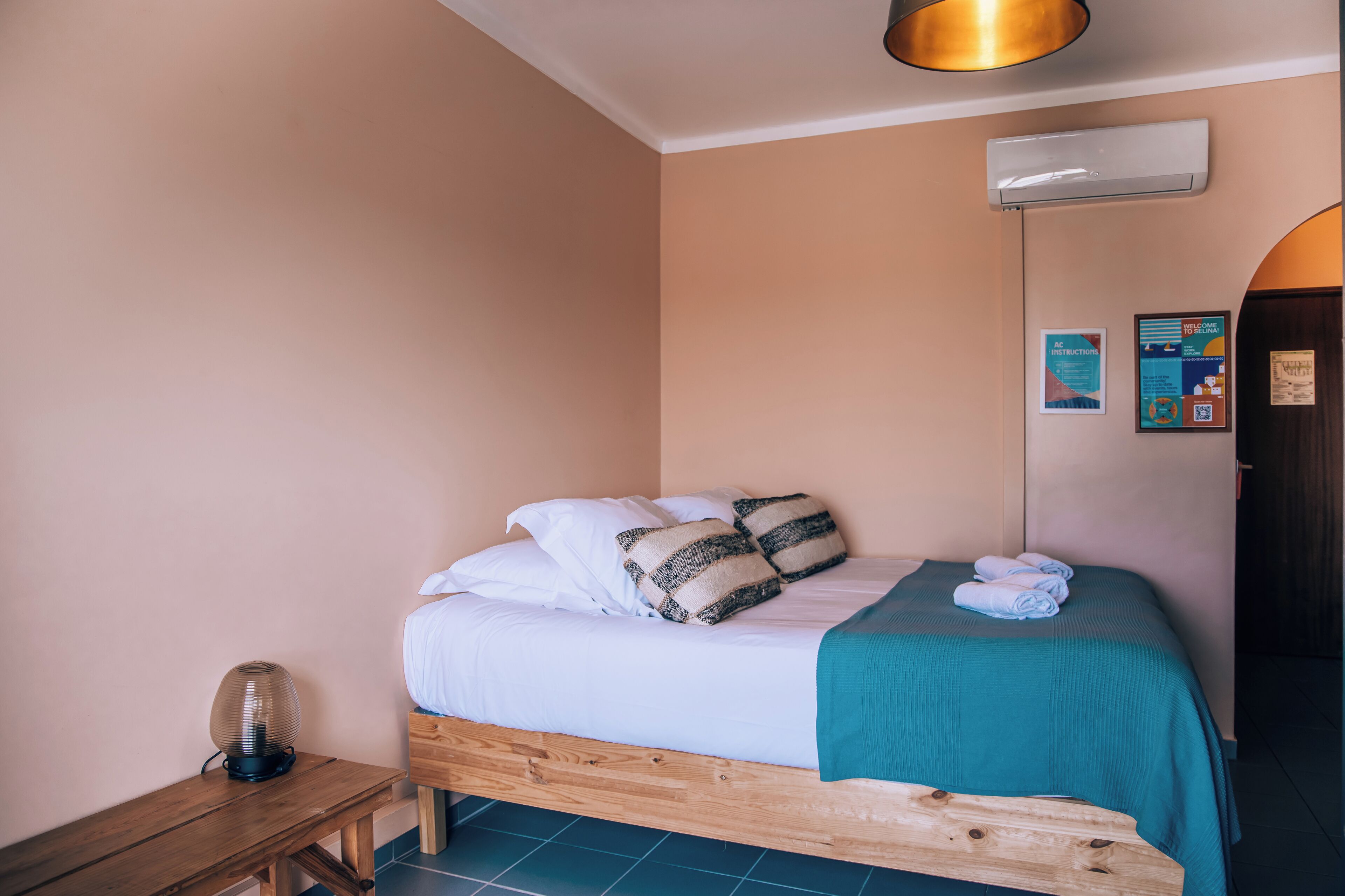 Un dormitorio con una cama de madera, ropa de cama blanca, un edredón verde azulado y una mesita de noche con una lámpara.
