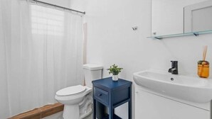 A bathroom with a toilet, a blue vanity with a plant, a glass shelf, and a white sink. - Otium | Walk to the Beach | Free Local Coffee (San Juan)