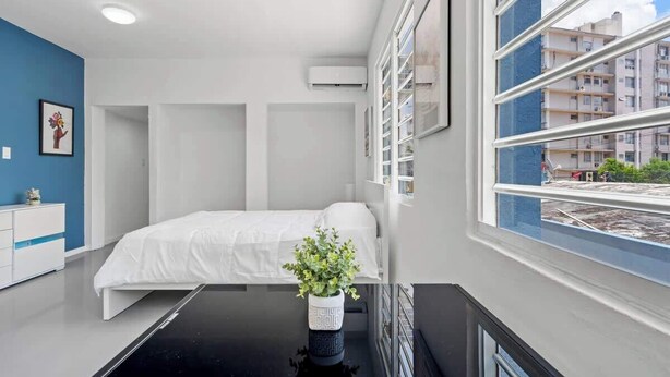 A modern hotel room with a bed, a small desk, a potted plant, and a window with a city view. - Otium | Walk to the Beach | Free Local Coffee (San Juan)