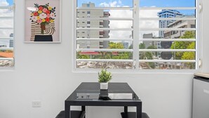 A room with a large window, a framed artwork of a bouquet, a small table with a potted plant, and a black chair. - Otium | Walk to the Beach | Free Local Coffee (San Juan)