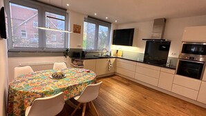 A modern kitchen with white cabinets, a black countertop, and stainless steel appliances. There is a dining table with a floral tablecloth and two chairs. - Vacation home in the Alter Hof - charming, central, with garden (Bad Zwischenahn)