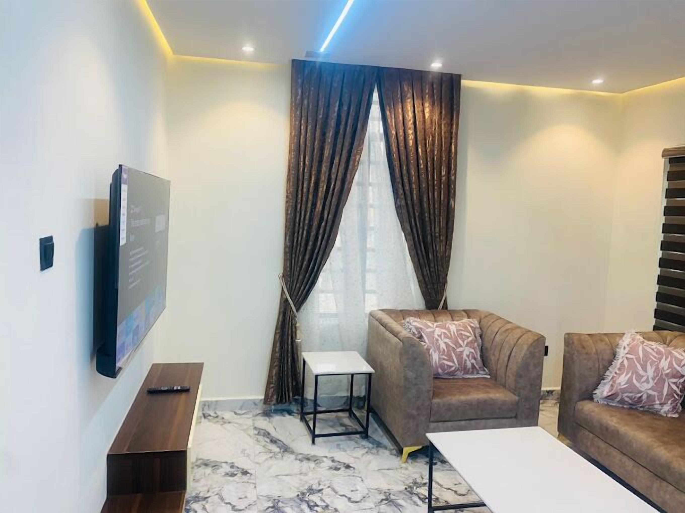Een moderne woonkamer met een flatscreen televisie aan de muur, een bank met gekleurde kussens, een klein wit bijzettafeltje en een vloer met marmerpatroon.