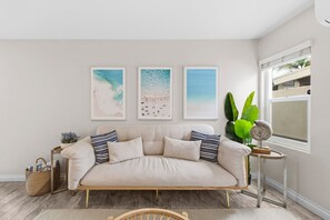 Sala de estar em tons neutros, com um sofá bege, uma pequena mesa lateral com uma planta e três quadros com tema de praia na parede.