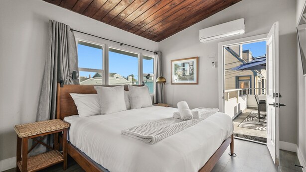Un dormitorio con techo de madera, una cama con ropa blanca, una mesita de noche y vistas al exterior a través de una puerta abierta.