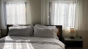 Ein ordentlich bezogenes Bett mit weißen Leinen, ein Nachttisch mit Lampe und durchscheinende Vorhänge an zwei Fenstern.