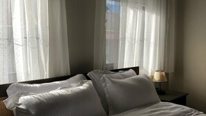 Ein Bett mit weißer Bettwäsche, ein Nachttisch mit Lampe und durchsichtige Vorhänge an einem Fenster.