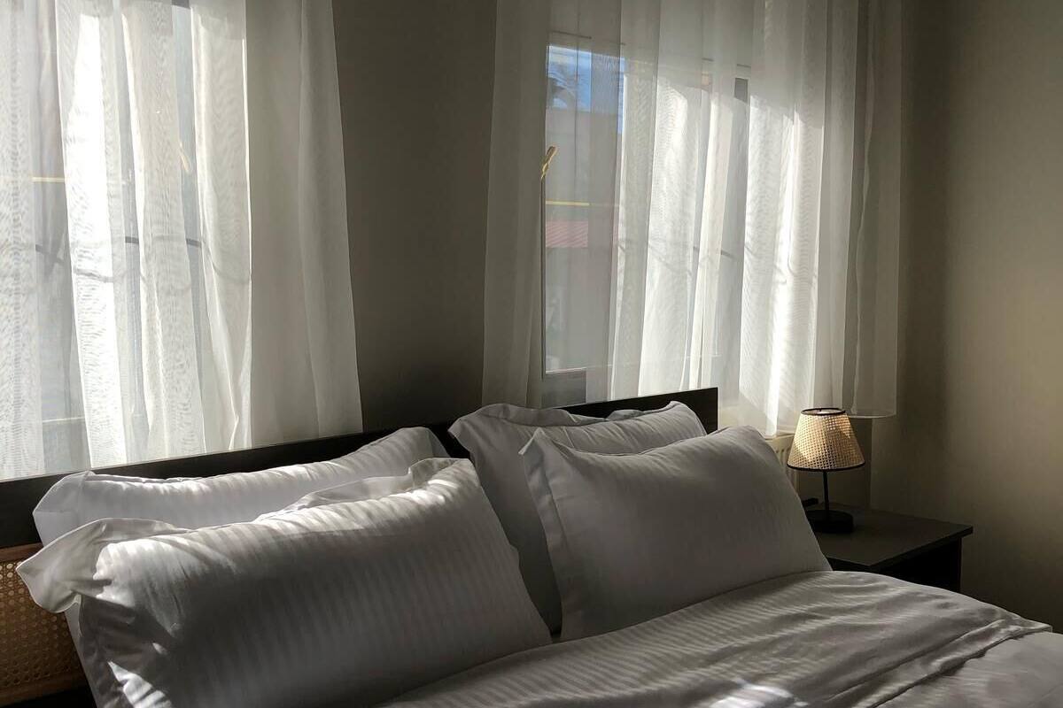 A bed with white bedding, a nightstand with a lamp, and sheer curtains on a window.