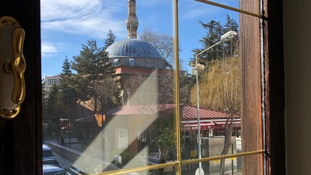 Blick durch ein Fenster mit Metallgitter, zeigt ein Gebäude mit Kuppel und Minarett, Bäume und eine Straßenszene.