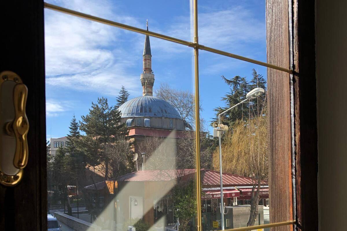 A view through a window with a metal grid, featuring a building with a dome and a minaret, trees, and a street scene.