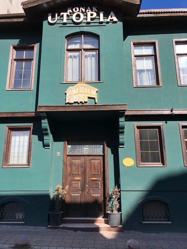 A green building with a wooden double door, potted plants, and a sign above the door. - Utopia Konak Otel (Bozuyuk)