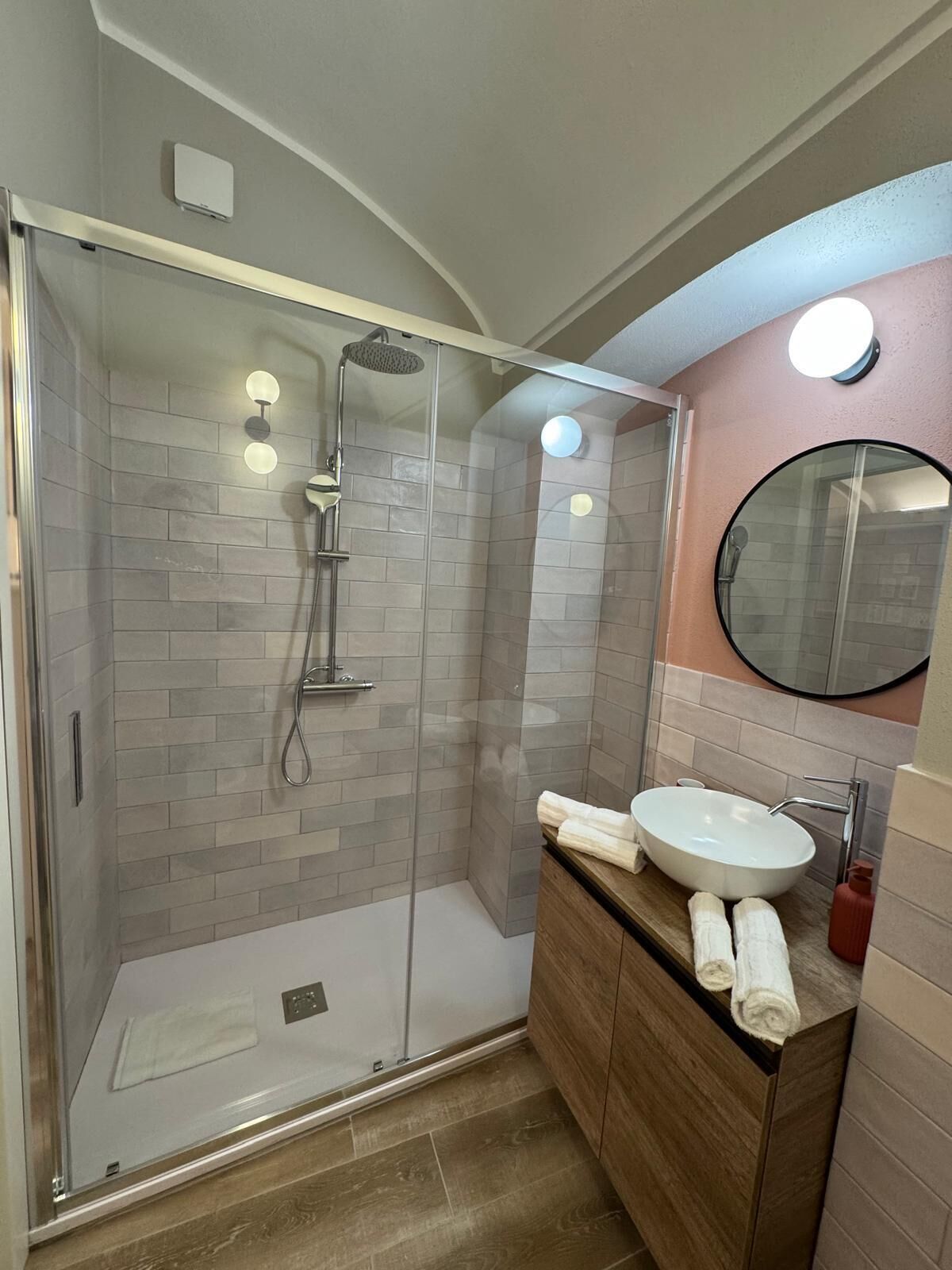Une salle de bain moderne avec une douche à parois de verre, un miroir rond et un meuble-lavabo en bois avec un vasque blanche.