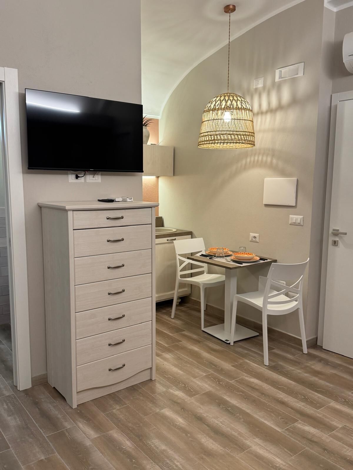 Une cuisine moderne équipée d’un téléviseur à écran plat, d’un meuble blanc avec des tiroirs, d’une table à manger avec des chaises blanches et d’un luminaire suspendu.