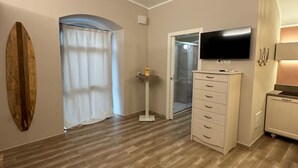 Una habitación amplia con suelo de madera, un televisor de pantalla plana montado en la pared, un aparador blanco y un gran ventanal con cortinas.