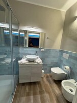 Un baño moderno con cabina de ducha de vidrio, tocador blanco con lavamanos y espejo, inodoro y bidé.