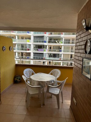 En balkon med et rundt bord og stole, flisegulv og udsigt til en høj bygning med balkoner.