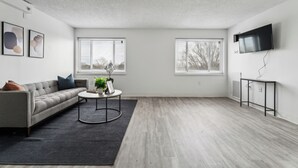 Een moderne woonkamer met een grijze bank, een ronde salontafel en een zwart vloerkleed. Er zijn twee ramen met zonwering, een flatscreentelevisie aan de muur en een radiator.