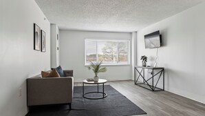 Een moderne woonkamer met een grijze bank, een ronde salontafel, een potplant en een aan de muur bevestigde televisie.