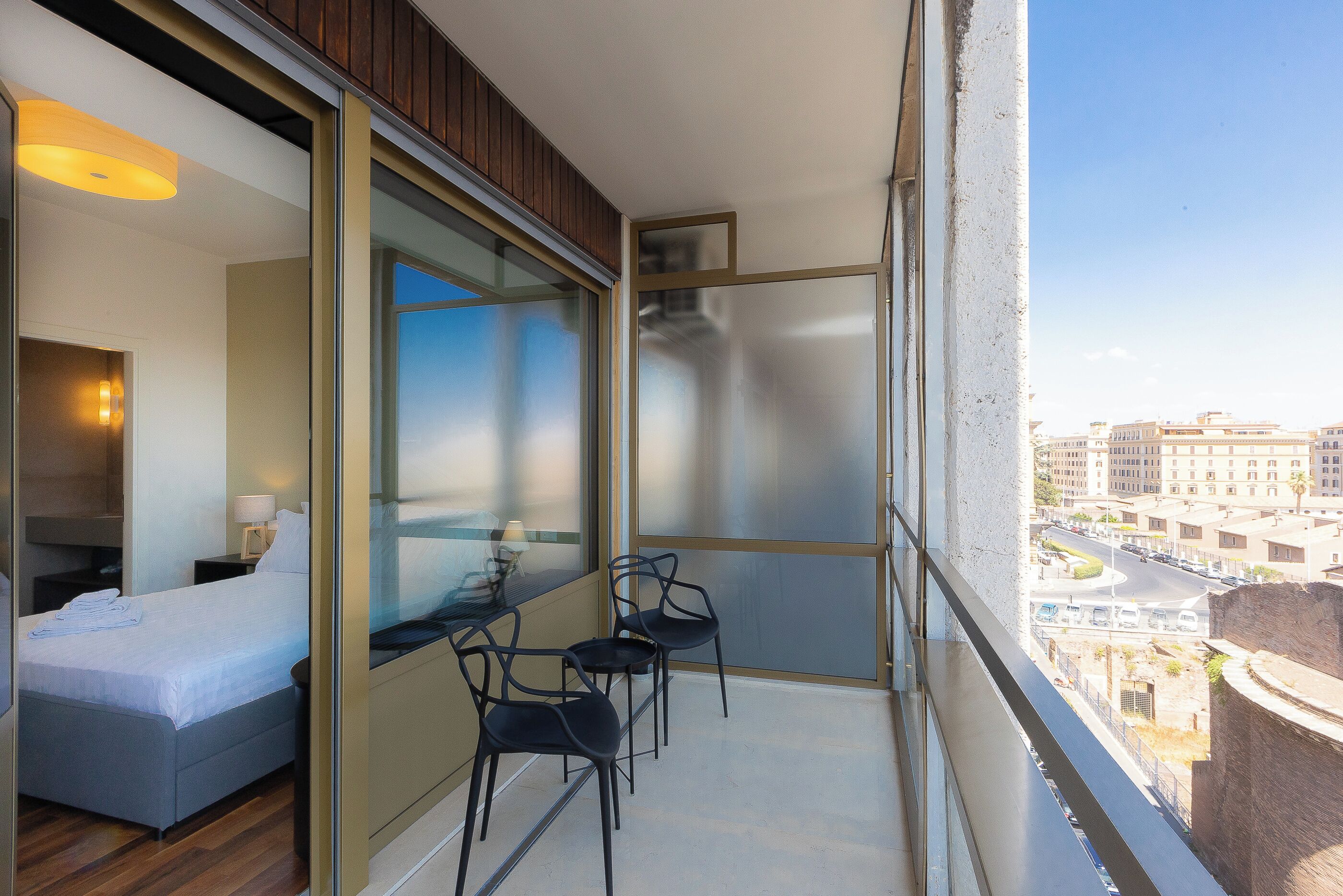 Un balcón con una cama, dos sillas y un muro de cristal, con vistas a la ciudad.