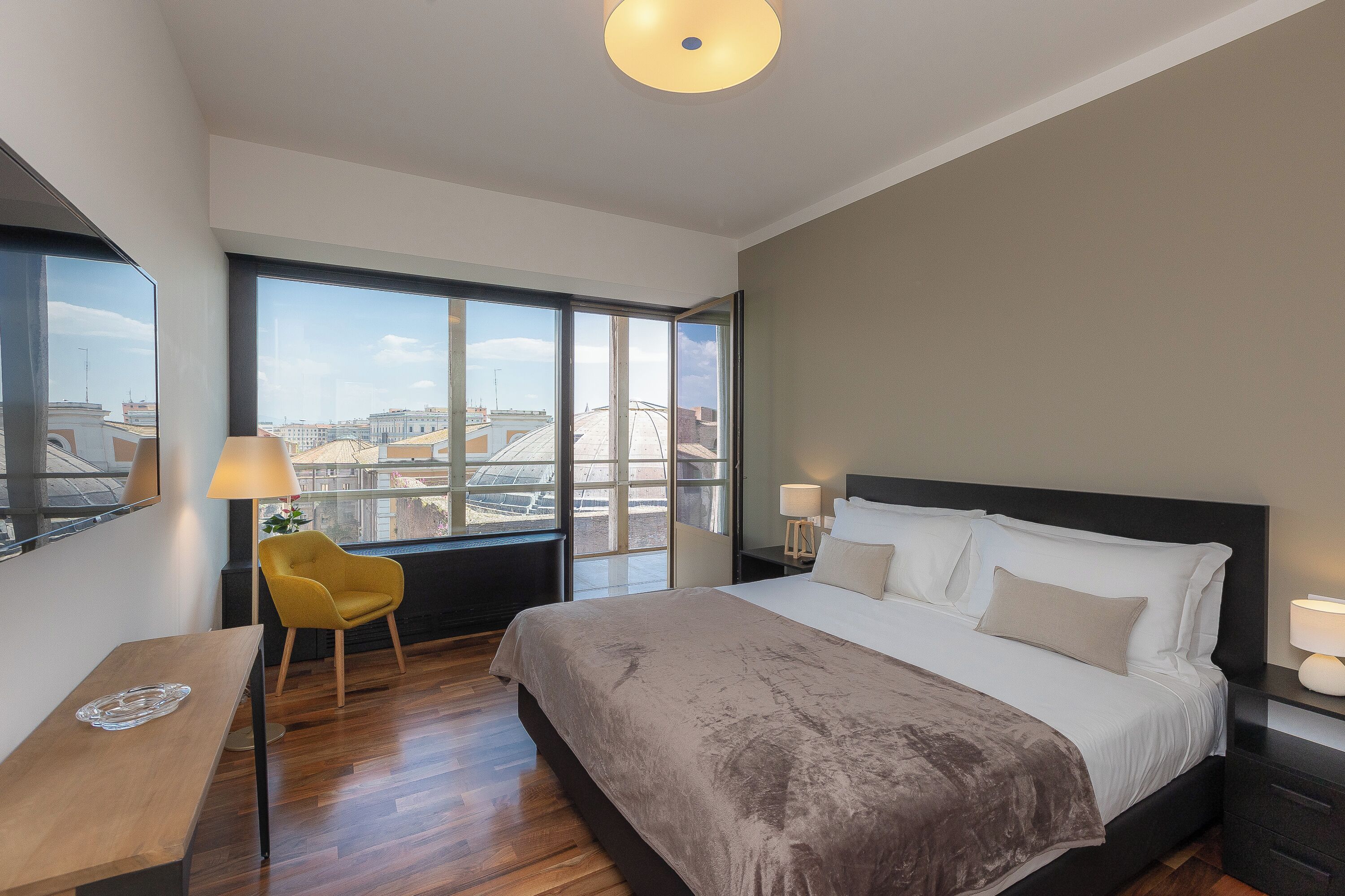 A modern hotel room with a large bed, a desk, a chair, and a view of the city through large windows.