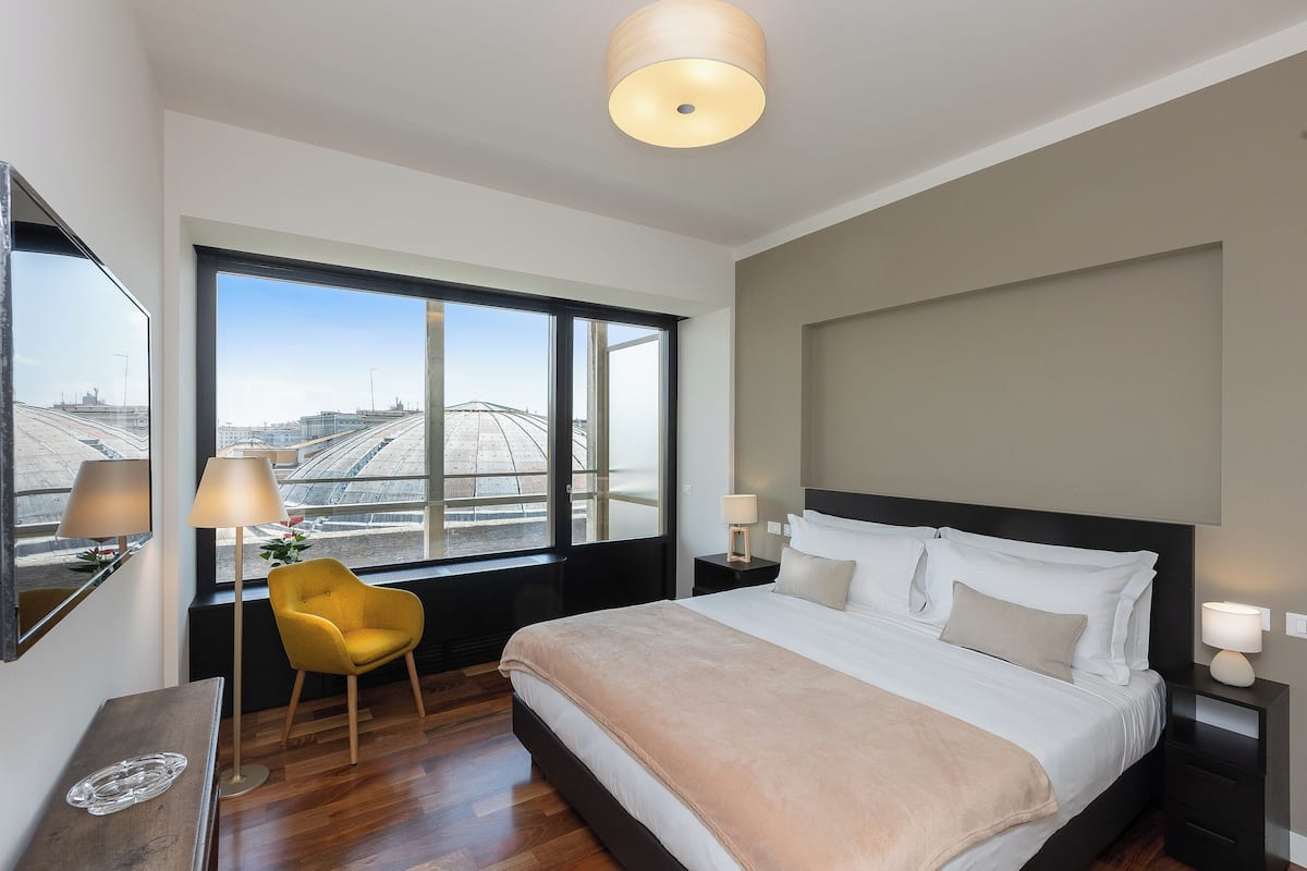 Ein modernes Hotelzimmer mit einem großen Bett, einem Sessel, einem Schreibtisch und Blick auf die Stadt durch große Fenster.