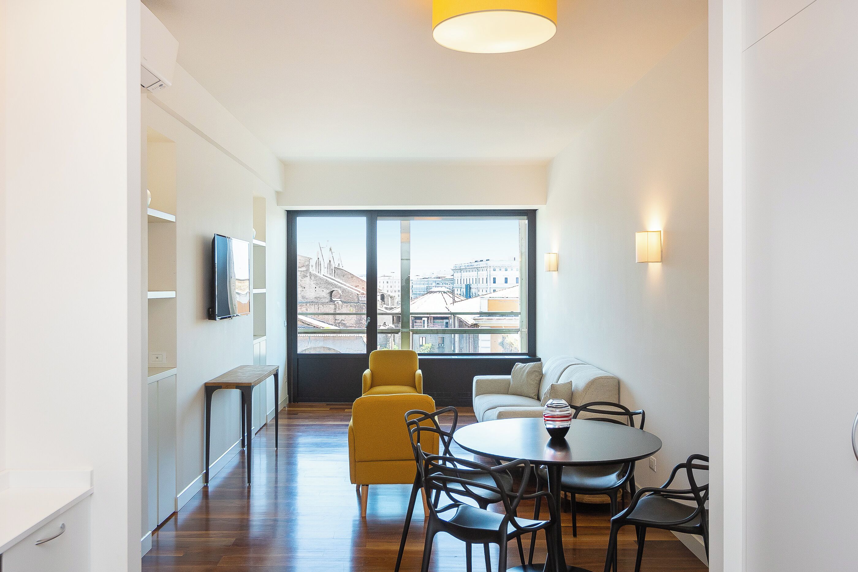 A modern living room with a sofa, yellow armchair, dining table, and chairs. A TV is mounted on the wall, and there is a large window with a view of buildings.