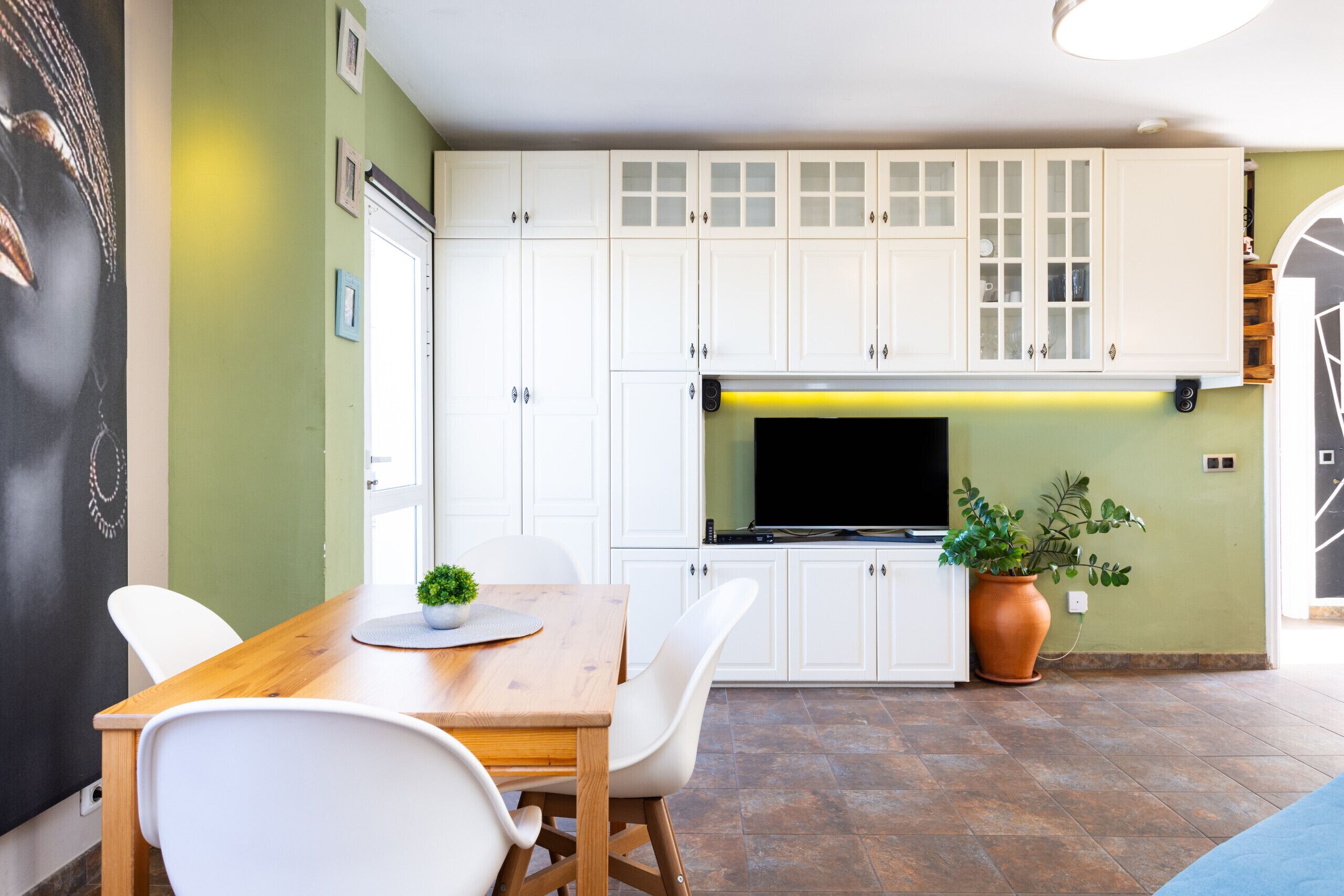 A modern kitchen with white cabinets, a wooden dining table, and a potted plant.