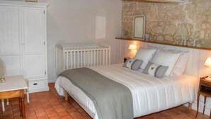 A bedroom with a stone wall, a white crib, a bed with pillows, a nightstand, and a small table with a lamp. - Charming Touraine cottage with spa and private garden (Saint-Germain-sur-Vienne)