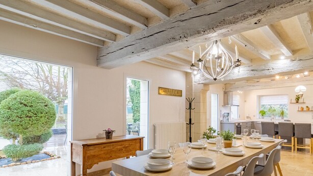 Un vaste espace dédié à la salle à manger, avec une longue table en bois pouvant accueillir six personnes, entourée de chaises modernes. Un lustre est suspendu au plafond et la pièce est agrémentée de poutres apparentes.