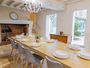 Ein Essbereich mit einem langen Holztisch, gedeckt für sechs Personen, umgeben von grauen Stühlen. Eine Chandelüster hängt von der Decke, und im Hintergrund ist ein Kamin aus Ziegelsteinen zu sehen.