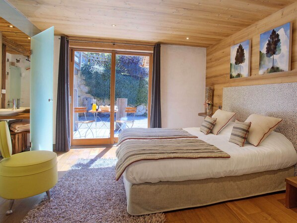 A bedroom with a large bed, a wooden headboard, and a view through a glass door to an outdoor seating area. - 5★ Chalet with spa, sauna & hammam for 15 people, close to slopes (La Clusaz)