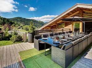 Un espacio de comedor al aire libre con una mesa larga preparada para varios invitados, rodeado de una terraza de madera y vistas a una cadena montañosa boscosa.